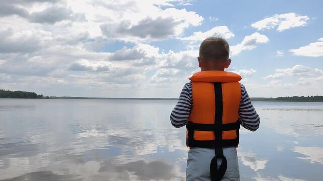 A young boy in bright orange life jacket stands facing vast, tranquil water body under partly cloudy sky serene landscape reflecting quiet wonder childhood exploration and the peaceful beauty nature