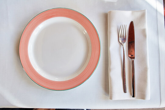 Top-down view of an empty coral-rim plate beside a neatly folded napkin with fork and knife on a white textured tablecloth, conveying dining readiness, minimalism and table etiquette.