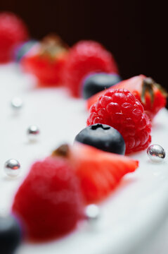 Close-up of a fresh berry cake decoration featuring raspberries, blueberries and sliced strawberries on smooth white cream, accented by silver dragees for an elegant dessert presentation.