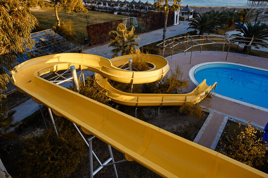 An empty yellow waterslide winds above a quiet blue pool at a closed outdoor waterpark by the beach, evoking off‑season stillness, summer memories, and urban leisure architecture in muted light.