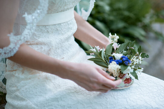 Bride wearing lace wedding dress delicately holds a small floral arrangement presented in a vintage teacup, evoking romantic, intimate, and vintage wedding styling with soft natural light and detail.