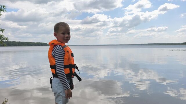A toddler in a bright orange life jacket stands by the calm water, highlighting essential child water safety and the peace of mind that proper gear brings to family outings near lakes and rivers.