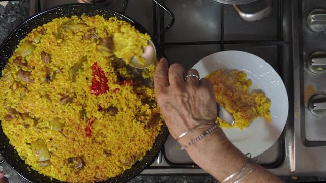 top view of an elderly woman hands serving delicious freshly cooked traditional spanish saffron rice paella from a large black pan onto a white ceramic plate in kitchen