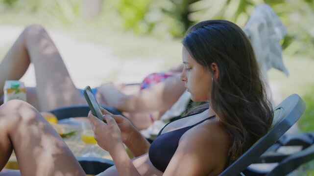 A teen woman holding a smartphone while sunbathing on a lounger by the pool with another woman.