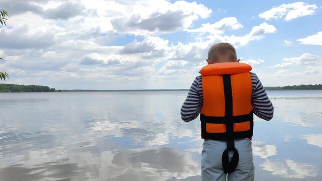 A young boy in bright orange life jacket stands facing vast, tranquil water body under partly cloudy sky serene landscape reflecting quiet wonder childhood exploration and the peaceful beauty nature