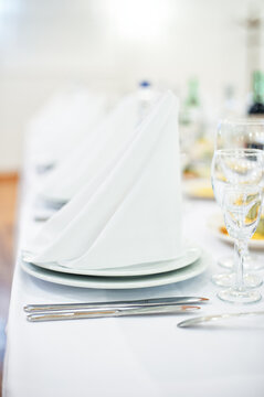 Elegant white place settings featuring folded napkins, stacked plates, silver cutlery and sparkling glassware on a linen-covered table for a wedding reception, formal banquet or upscale catered event.