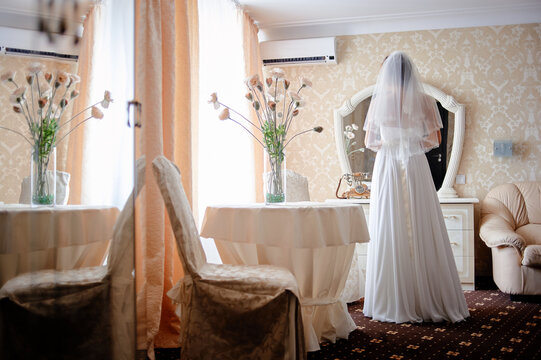 A bride wearing a flowing white gown and veil stands at a vintage vanity mirror in an elegant hotel room, bathed in soft light with floral arrangements and quiet anticipation before the ceremony.