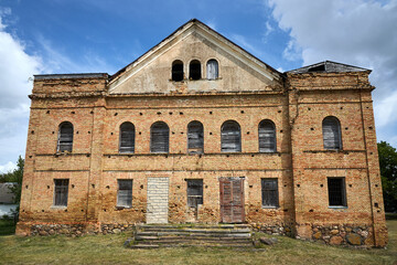 Fototapeta premium Abandoned Indura Synagogue in Grodno region, Belarus, monumental red brick building. Classic architectural structure features symmetry, arched windows, and triangular pediment under cloudy blue sky.