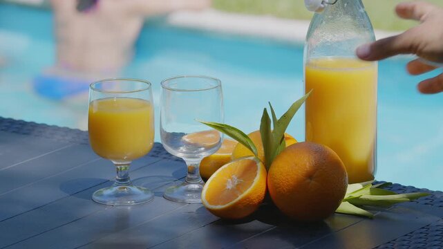 A man pours orange juice into a glass on a poolside table with blurred background of two young women sitting on the edge of the pool.