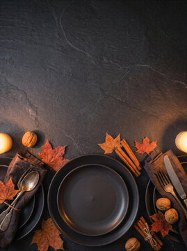 Elegant autumn table setting with black plates, napkins, maple leaves, walnuts and candles on a dark stone background with copy space. Thanksgiving dinner concept.