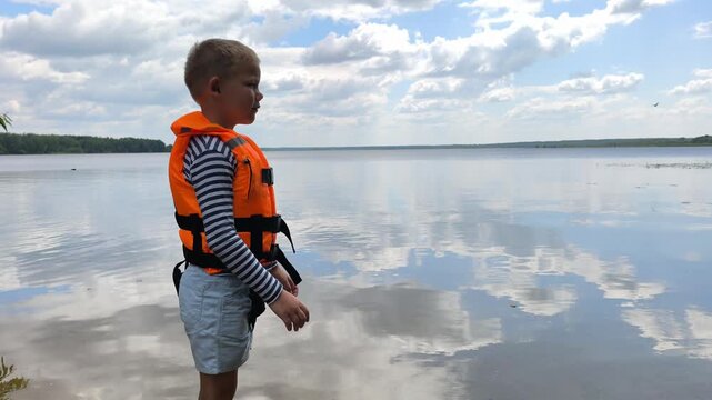 A toddler in a bright orange life jacket stands by the calm water, highlighting essential child water safety and the peace of mind that proper gear brings to family outings near lakes and rivers.
