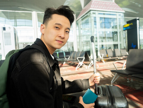 a young man wearing a black jacket sits in a bright airport waiting area, looking back at the camera while holding a blue passport and his suitcase