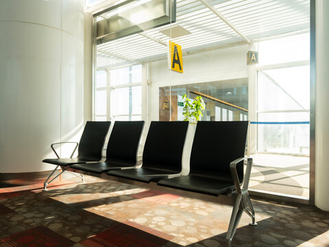 a row of four empty black waiting chairs sits on a patterned carpet in a brightly sunlit airport terminal beneath a yellow "A" sign