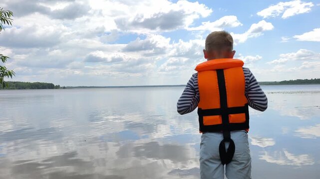 A young boy in bright orange life jacket stands facing vast, tranquil water body under partly cloudy sky serene landscape reflecting quiet wonder childhood exploration and the peaceful beauty nature