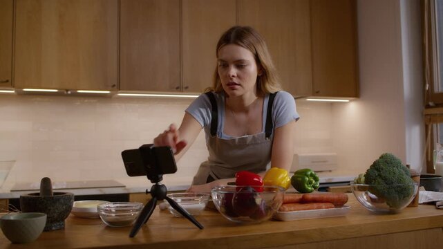 Woman leaning on the kitchen counter with a variety of vegetables broccoli and colorful bell peppers laid out on the surface in preparation for making a salad. Looking into a mobile phone for recipe