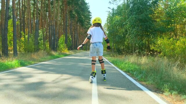 Child girl in casual clothes roller skating on road in forest on sunny summer day.