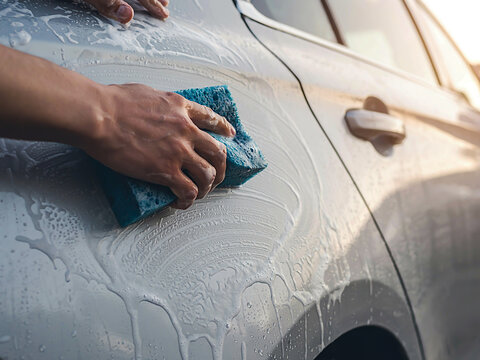 Close-up photo of a man washing a silver car door with blue sponge and soapy foam during vehicle detailing and auto maintenance service