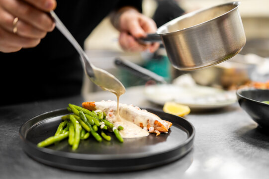 A chef in a commercial kitchen plates a dish, pouring a creamy white sauce from a small metal pot over a grilled salmon fillet served with green beans on a sleek black plate