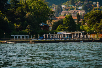 Obraz premium Lakeside resort with sun loungers and umbrellas along the shore, surrounded by trees and buildings in a picturesque setting