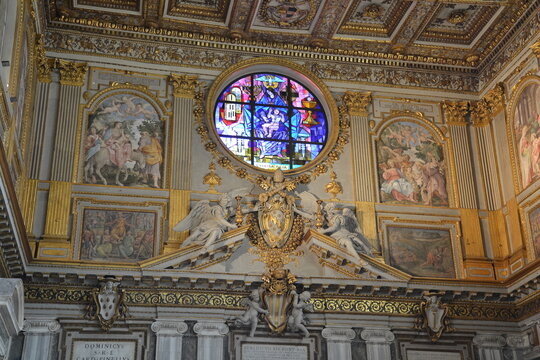 Stained glass rose window, gold coat of arms with angels, and frescoes inside Basilica di Santa Maria Maggiore, Rome. Ornate Renaissance and Baroque interior details.