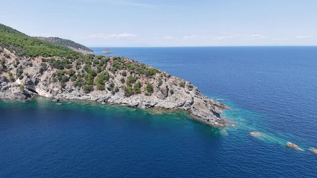High-angle drone view gradually reveals a rugged forested peninsula surrounded by clear shallows and deep blue sea.