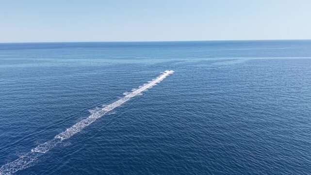 A small distant watercraft travels across expansive blue water, producing a long foamy trail before beginning a gentle turn.