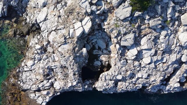 Aerial pullback expands from fractured coastal rocks and dark cavities to a wider view of a forested shore and transparent blue sea.