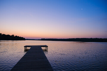 Fototapeta premium Moscow. Russia. 25.07.2026 Wooden pier extending into a calm lake at dusk. Serene sunset landscape with moonlight and twilight sky over water. Peaceful nature scene for travel and relaxation concept b