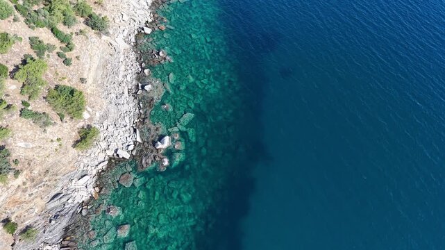 Aerial view moves along a rugged rocky shoreline bordered by transparent coastal water and darker open sea.