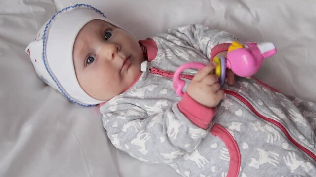 Cute newborn baby lying in bed and holding a pink rattle toy close up, Little baby boy or girl holding a colorful toy rattle in a crib