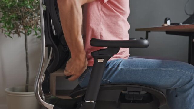 Man sits in an ergonomic office chair, adjusts the lumbar support lever for better posture during computer work
