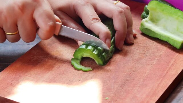 Close-up of a woman's hands slicing fresh bitter melon on a wooden cutting board while preparing a healthy homemade meal. Home cooking, vegetable preparation, and nutritious food.