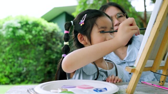 Happy Asian mother and young daughter painting on canvas together in the garden, cute little girl enjoying creative art workshop activity with mom
