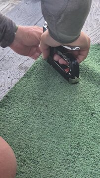 Stapling, artificial turf, hand securing green outdoor carpet onto a rustic wooden deck during a home improvement project. Vertical video.