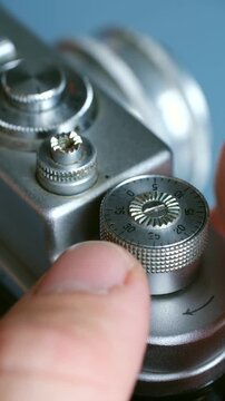 Close up photographer thumb turning mechanical self timer dial on classic silver analog camera, preparing to take picture with delayed shutter release. Vertical. Man use vintage analog camera settings