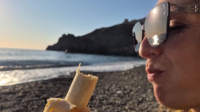 Young woman eating banana on a rocky beach at sunset. Close-up static side profile shot.