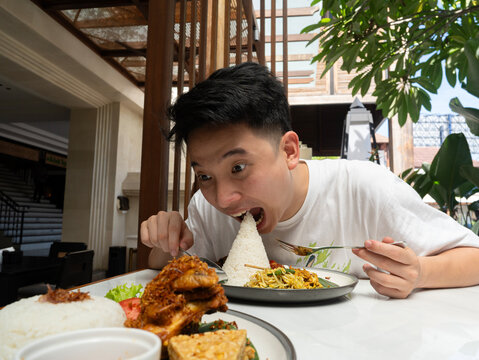 A young man makes a playful, wide-eyed expression as he leans in to pretend to take a massive bite out of a cone of rice at a sunny, semi-outdoor restaurant