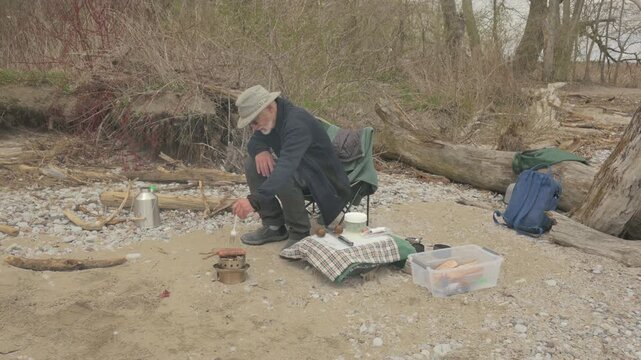 Two senior men react as sausage falls from camp grill onto beach sand at lakeshore picnic site, Lynde Shores Conservation Area, Whitby, Ontario, Canada.