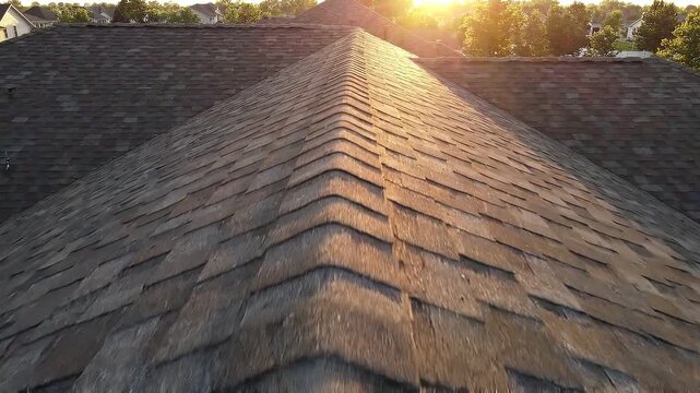 Asphalt shingle roof ridge seen from above during sunset. Aerial drone view of residential building exterior surface. Construction maintenance and housing concept.