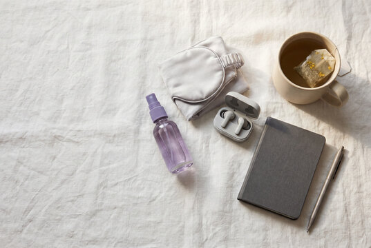A minimal flat lay of self-care and sleep essentials on white linen including a sleep mask, wireless earbuds, lavender spray, a gray notebook, pen, and herbal tea.
