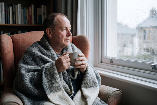 A middle-aged man wrapped in a gray plaid blanket holds a mug and gazes thoughtfully out a rain-streaked window from a comfortable armchair beside a bookshelf.
