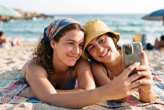 Two young women lying on a colorful beach towel smile and take a selfie together with the ocean in the background.
