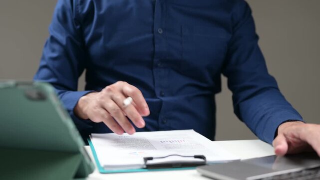 Male entrepreneur analyzes corporate report paper sheet inside clipboard, holding digital stylus pointer near touch screen tablet, typing laptop keyboard keys inside workspace desk environment.