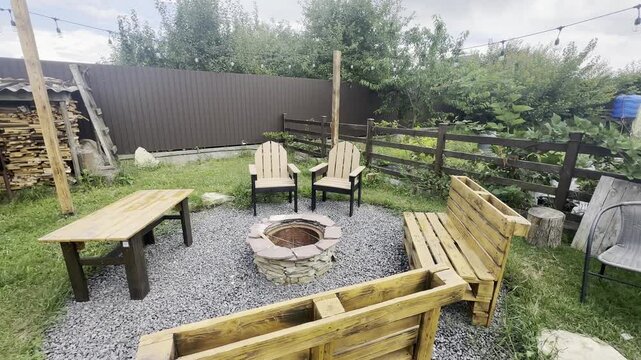 Wooden seating surrounds stone fire pit in landscaped backyard with gravel, grass, fence, trees, and rustic furniture under natural daylight.