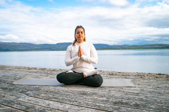 Woman meditating in lotus position with prayer hands on a wooden pier by a scenic fjord in Norway