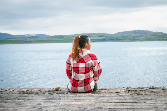 Rear view of a young woman sitting on a wooden pier looking at a scenic fjord in Norway