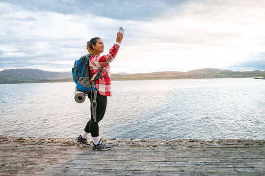 Female backpacker standing on a wooden pier taking a selfie or photo with smartphone by the fjord in Norway