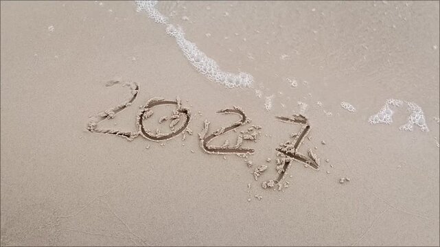 2027 handwritten in sand on beach with sea wave foam approaching.