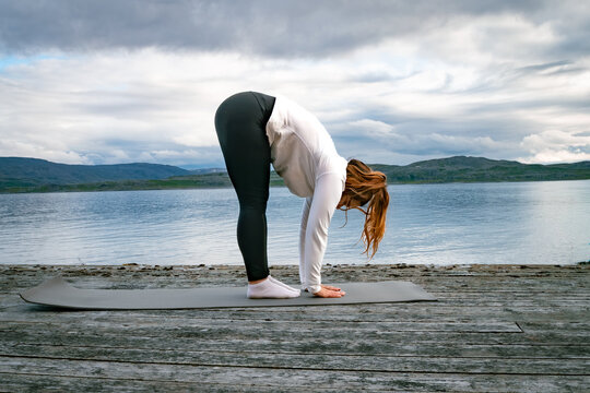 Woman practicing standing forward fold yoga pose on a mat at a wooden pier by the fjord in Norway