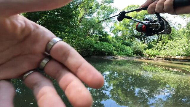 POV angler casting spinning rod in a wild forest stream. Close-up on hands and fishing reel during summer fishing.